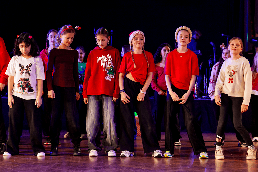 Kinder in weihnachtlicher Kleidung mit Pullovern und Mützen tanzen auf einer Bühne bei einer Aufführung