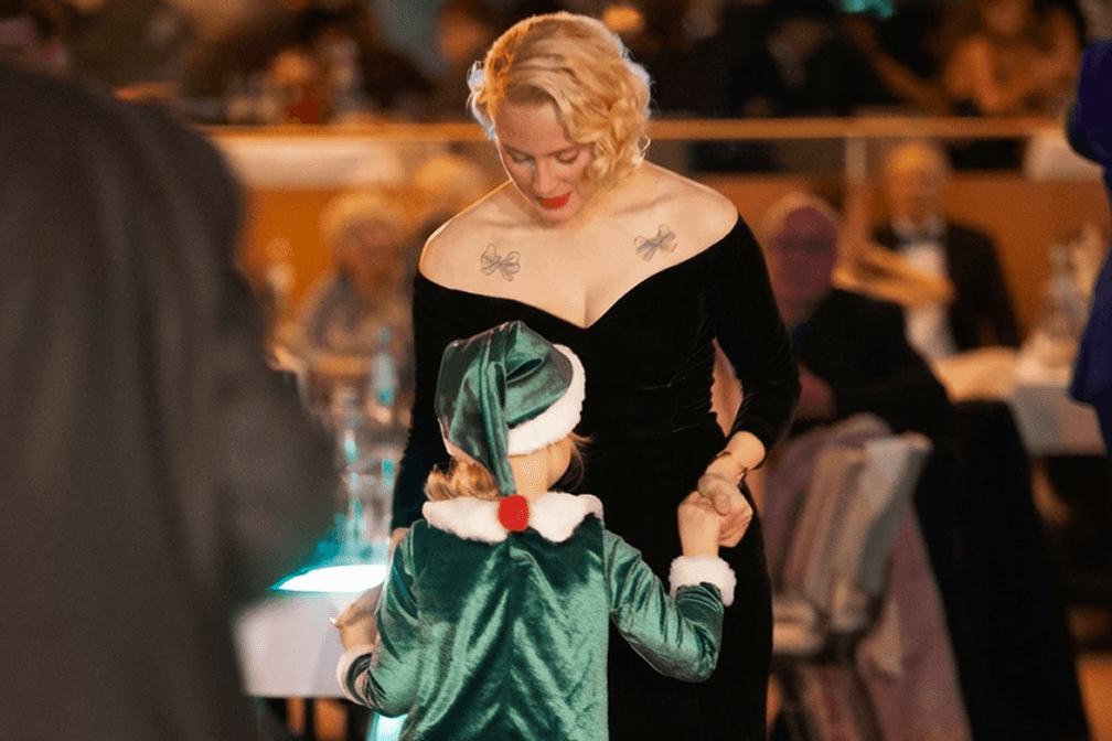 Blonde Frau im schwarzen Samtkleid hält Händchen mit Kind im grünen Samt-Weihnachtskostüm bei Veranstaltung