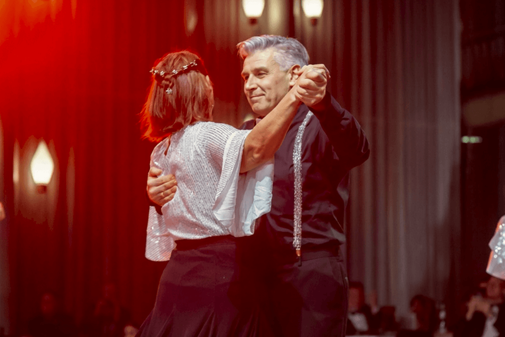 Älteres Paar tanzt in elegantem Saal bei warmem Licht Mann in schwarzem Hemd und Frau in weißer Bluse mit Haarreif