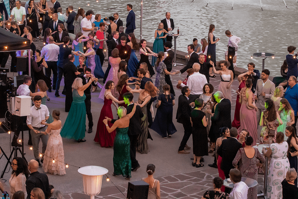 Tanzende Menschen in festlicher Kleidung bei einer Abendveranstaltung am Wasser mit Lichterketten und Stehtischen