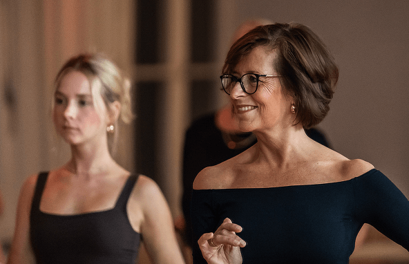 Zwei Frauen bei Tanzunterricht eine trägt schwarze Brille und schwarzes schulterfreies Oberteil, helle Holzwände im Hintergrund