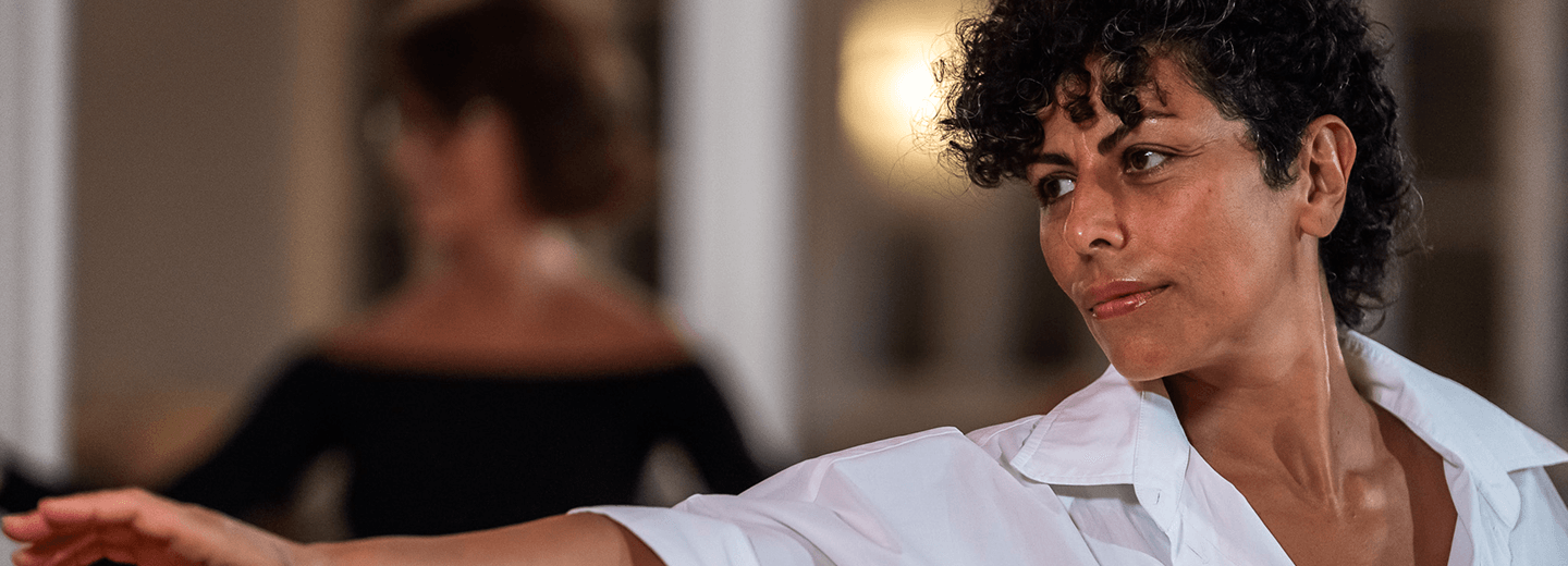 Frau mit lockigem Haar und weißem Hemd beim Tanz in hellem Raum mit Spiegel im Hintergrund