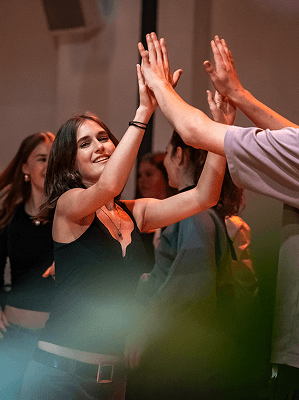 Junge Frau in schwarzem Oberteil gibt High Five beim feierlichen Treffen in Innenraum mit anderen Menschen