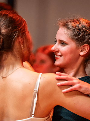 Zwei junge Frauen im Tanz, eine mit Zopf und schwarzem Oberteil, die andere in weißem Spaghettiträger-Top, warme Beleuchtung