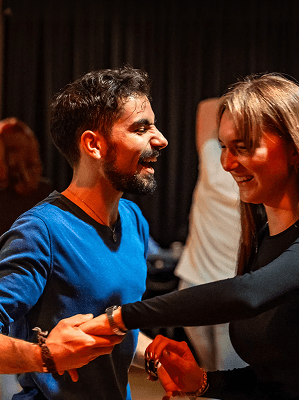 Zwei Personen beim Tanzen in einem Raum mit dunklem Hintergrund, beide lächeln und halten sich an den Händen