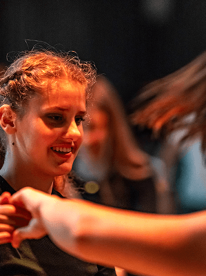 Junge Frau mit geflochtenem Haar tanzt und lächelt in einem dunklen Raum mit verschwommenen Personen im Hintergrund