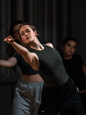Junge Frau in schwarzem Tanktop und lockerer Hose beim Tanztraining in Innenraum mit weiteren Tänzerinnen im Hintergrund