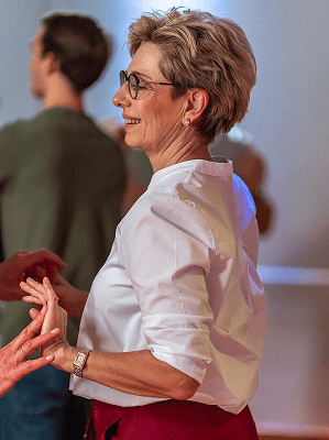 Ältere Frau mit Brille in weißer Bluse hält Hände einer anderen Person in freundlicher Umgebung