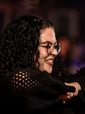 Frau mit schwarzen lockigen Haaren und Brille trägt schwarzen Pullover mit offenem Maschenmuster und lächelt in warmer Beleuchtung