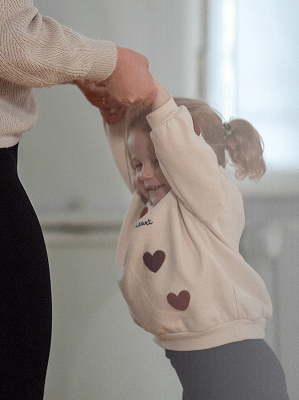 Kind mit hellbeigem Pullover mit Herzmotiven hält Hände eines Erwachsenen in hellem Raum