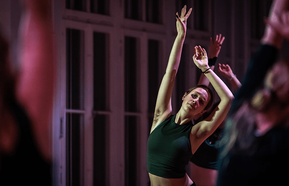 Frau im schwarzen Sporttop bei Dehnübung in hellem Raum mit großen Fenstern und anderen Teilnehmerinnen