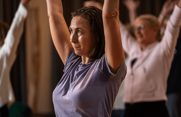 Frau in lila Shirt bei Yogaübung mit erhobenen Armen in hellem Raum mit weiteren Teilnehmern im Hintergrund