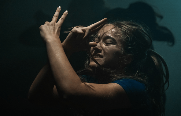 Junges Mädchen mit blonden Haaren und blauem Shirt macht mit Händen Gesten vor dunklem Hintergrund