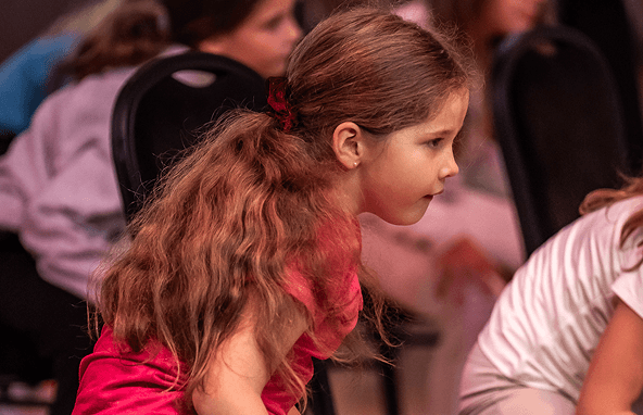 Mädchen mit roten Haaren und rotem Shirt sitzt konzentriert bei einer Veranstaltung mit weiteren Kindern im Hintergrund