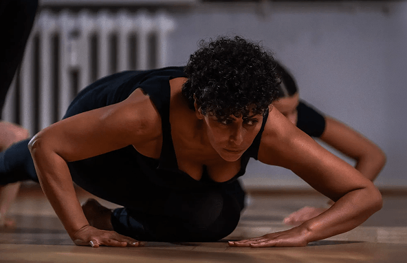 Frau mit dunklen Locken in schwarzer Sportkleidung bei Yoga-Übung auf Holzfußboden in Innenraum