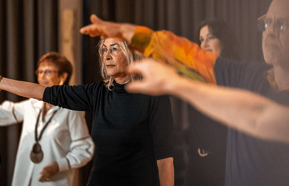 Ältere Menschen üben gemeinsam mit ausgestreckten Armen in einem Innenraum mit dunklem Vorhang