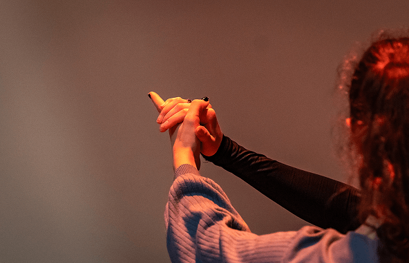 Zwei Hände in schwarzem und blauem Langarmshirt verschränkt vor braunem Hintergrund in warmem Licht