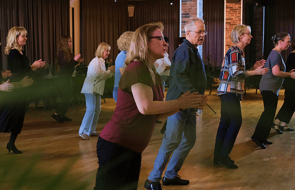 Gruppe älterer Menschen beim gemeinsamen Tanztraining in einem Saal mit Holzboden und Backsteinwand