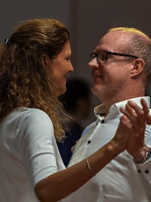 Paar mittleren Alters tanzt eng miteinander in heller Kleidung vor dunklem Hintergrund