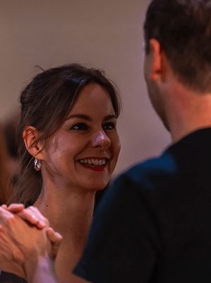 Frau mit braunen Haaren und silbernen Ohrringen lächelt einem Mann in schwarzem T-Shirt in gemütlicher Innenraumbeleuchtung zu