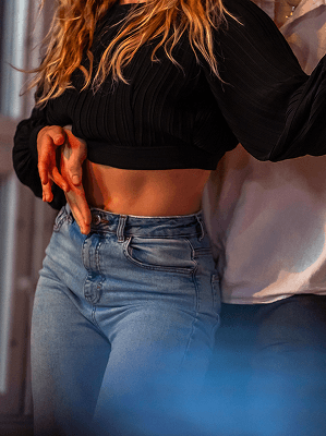 Frau in schwarzem Pullover und blauen Jeans mit barem Bauch beim Tanz mit Partner in Innenraum