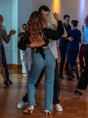 Junges Paar tanzt eng im Tanzsaal mit Holzfußboden und anderen tanzenden Paaren im Hintergrund