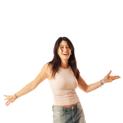 Frau mit langen dunklen Haaren in beigem Tanktop und blauen Jeans lacht und breitet die Arme aus vor weißem Hintergrund