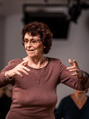 Ältere Frau mit braunem Pullover und Brille zeigt Gesten in einem Innenraum mit unscharfem Hintergrund