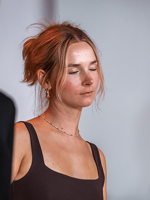 Frau mit roten Haaren und goldfarbener Kette und Ohrringen in schwarzem ärmellosen Oberteil mit geschlossenen Augen