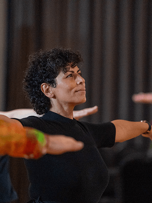 Frau mit kurzen lockigen Haaren in schwarzem Oberteil übt stehende Yoga-Pose in Innenraum mit Vorhängen