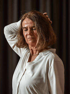 Frau mit mittellangen braunen Haaren und weißer Bluse hält Hand an den Kopf vor dunklem Hintergrund