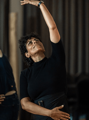 Frau mit schwarzen Locken in schwarzem Oberteil hebt einen Arm nach oben und blickt nach oben in Innenraum