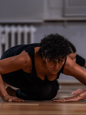 Frau mit lockigen dunklen Haaren in schwarzer Sportkleidung beim Yoga in einem Raum mit Holzboden und Heizkörper im Hintergrund