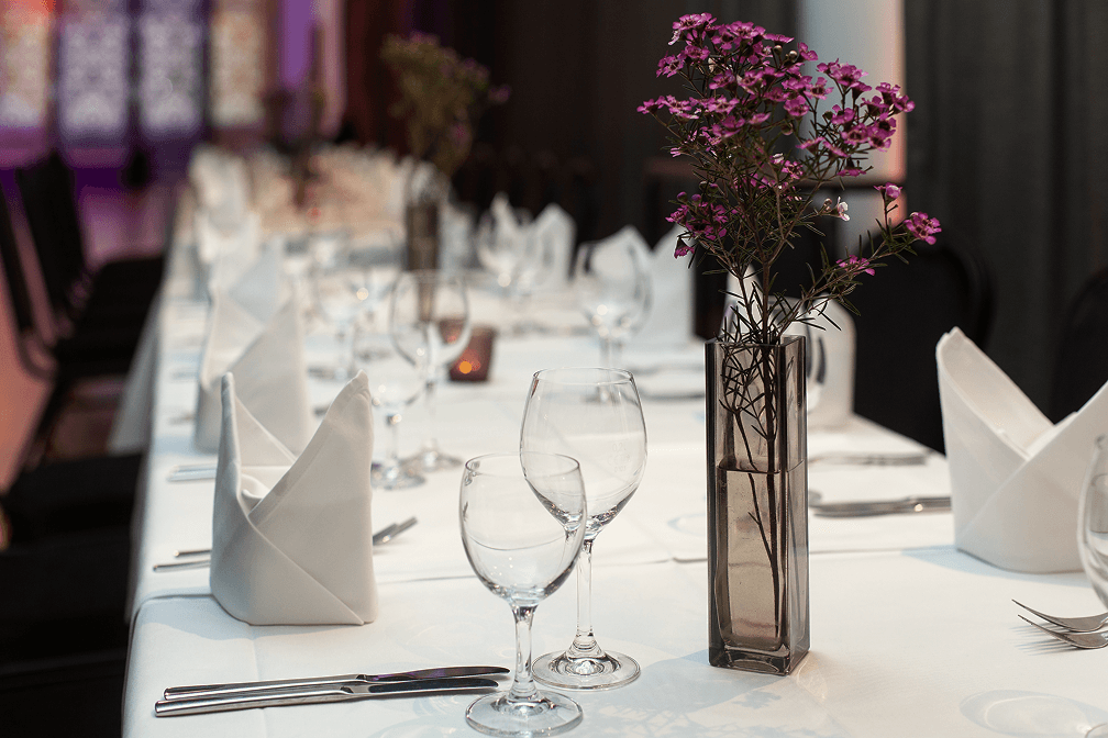 Festlich gedeckter Tisch mit weißen Stoffservietten, Gläsern und Vase mit lila Blumen in elegantem Ambiente