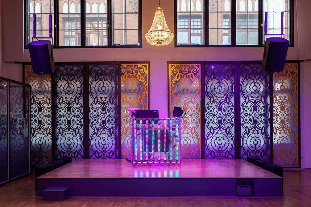 Leere Bühne mit moderner DJ-Konsole aus LED-Leisten vor verzierten Fenstern und Kronleuchter in heller Veranstaltungshalle