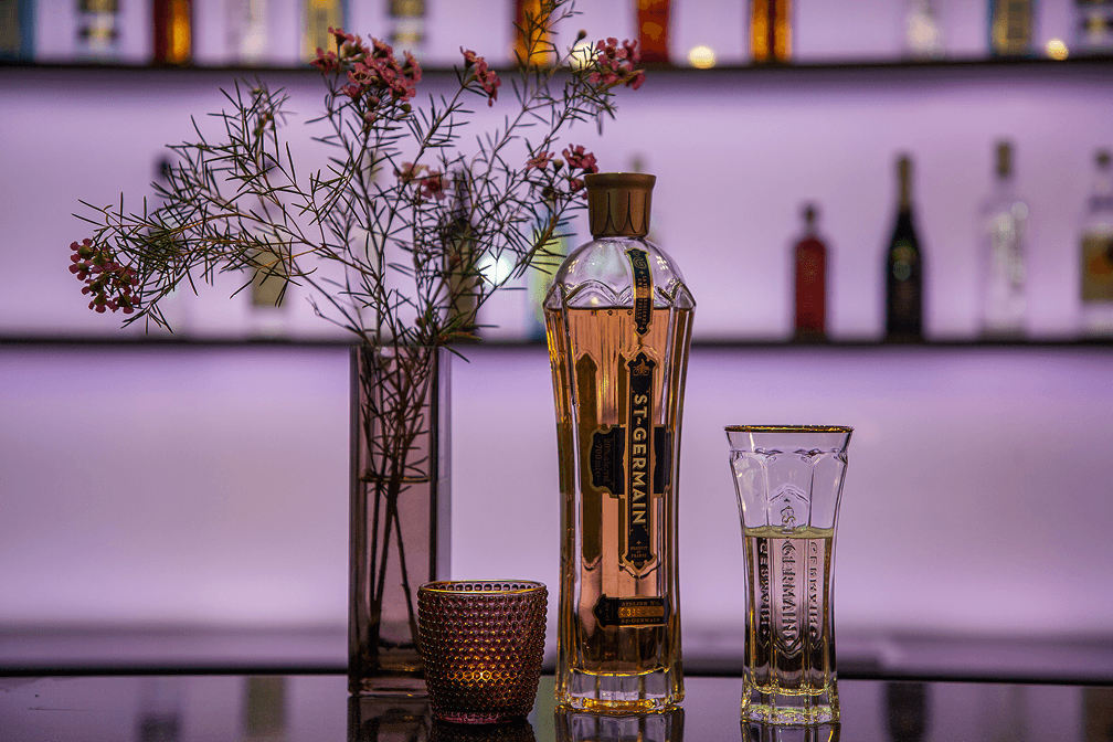 Flasche St. Germain Likör mit Glas und Vase mit kleinen rosa Blüten vor beleuchtetem Barregal