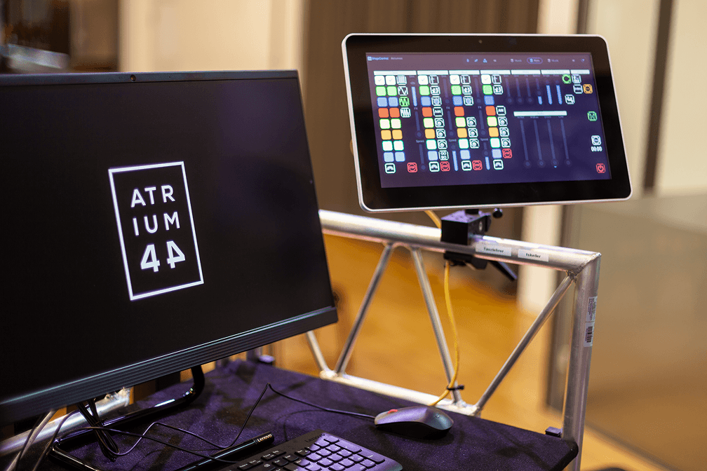 Computermonitor mit Atrium44 Logo neben Tablet mit farbigen Bedientasten auf Metallgestell in Innenraum