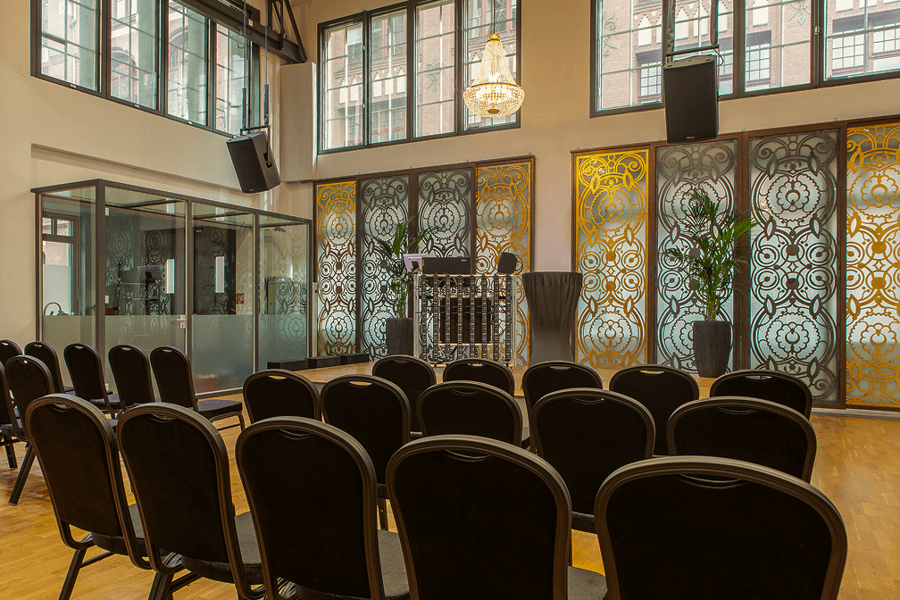 Moderner Veranstaltungsraum mit schwarzen Stühlen, Glaskabine, verzierten Fenstern und Kronleuchter in hellem Saal