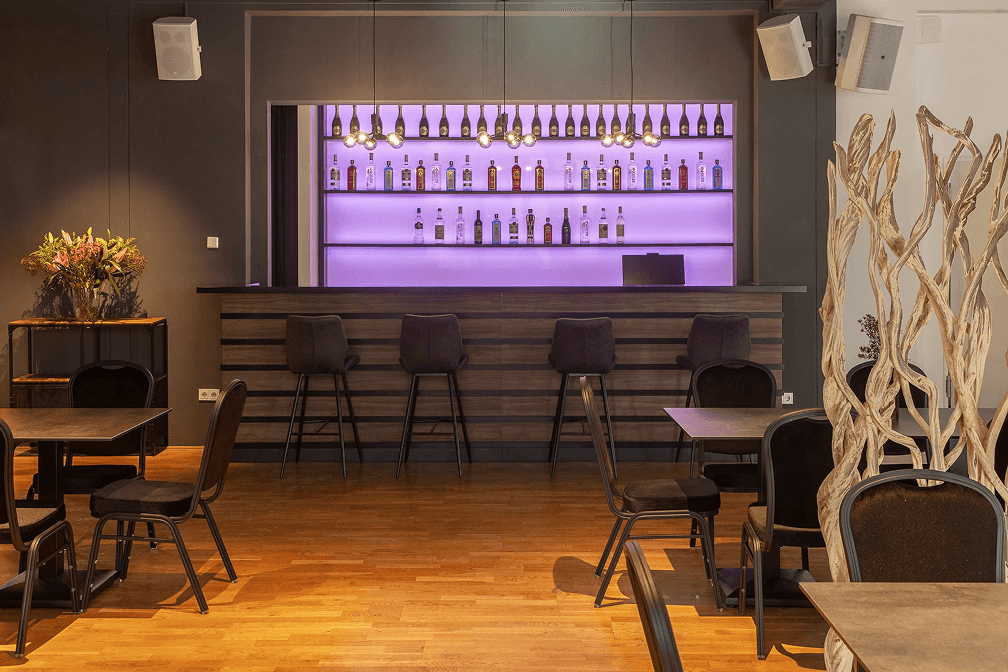 Moderne Bar mit Holztheke, schwarzen Barhockern, beleuchteten Regalen mit Spirituosen und Sitzbereich mit Tischen und Stühlen