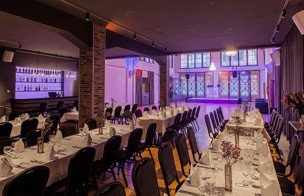 Festlich gedeckte Tische mit weißen Tischdecken und Blumenarrangements in einem stilvoll beleuchteten Veranstaltungsraum mit großer Fensterfront