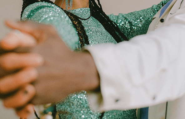 Zwei Personen halten sich an den Händen, eine trägt ein glitzerndes grünes Oberteil und lange Zöpfe