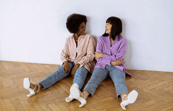 Zwei Frauen in bunten Hemden und Jeans sitzen entspannt auf Parkettboden vor weißer Wand und unterhalten sich