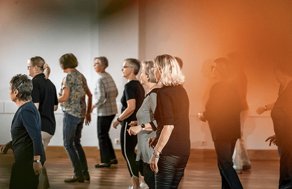 Gruppe älterer Menschen bei Tanzstunde in hellem Raum mit Holzboden und weißer Wand
