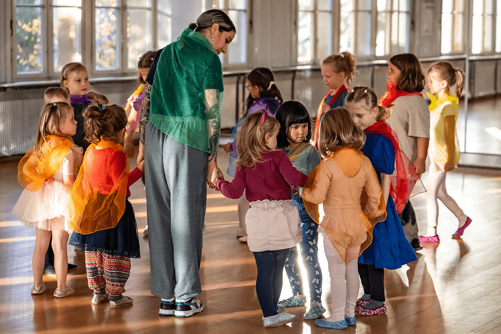 Tanzlehrerin steht mit einer Gruppe kleiner Kinder im Kreis in einem hellen Tanzsaal und hält zwei Kinder an der Hand, während die Kinder bunte Tücher tragen.