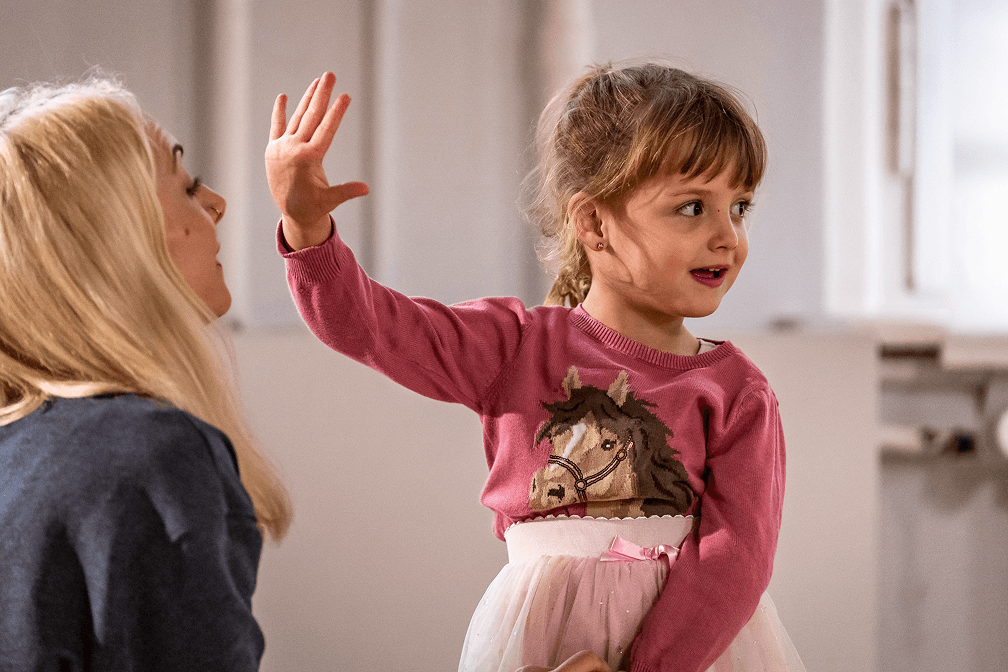 Kleines Mädchen im Tutu hebt lächelnd die Hand im Tanzunterricht, neben ihr sitzt eine erwachsene Begleitperson im Tanzsaal