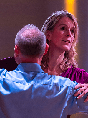 Paar beim Tanzen in Innenraum mit warmem Licht, Frau in lila Oberteil, Mann in blauem Hemd von hinten zu sehen