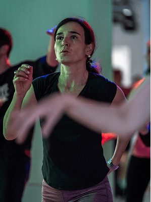 Frau mit dunklem Haar und schwarzem Oberteil bei Tanz- oder Fitnesskurs in Innenraum mit Gruppenbeteiligung