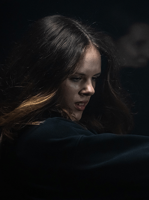 Frau mit langen braunen Haaren und nachdenklichem Blick vor dunklem Hintergrund in Innenraum