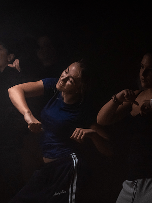 Junge Frau in blauem T-Shirt tanzt energiegeladen in dunkler Umgebung mit anderen Personen im Hintergrund