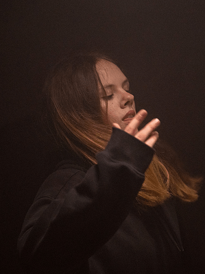 Junge Frau mit braunen Haaren in schwarzem Pullover hebt Hand vor dunklem Hintergrund